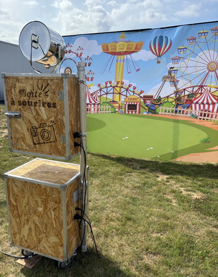 La borne « Boîte à sourires » en bois recyclé, avec son projecteur, devant un fond fête foraine en extérieur